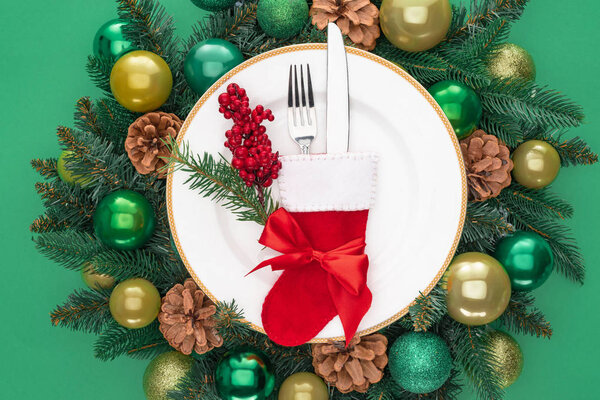 view from above of fork and knife in santa sock and red berries on plate surrounded by evergreen tree branches with christmas balls isolated on green