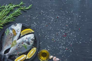 view from above of raw fish with lemon, bay leaves and rosemary in tray on black table covered by salt and pepper clipart