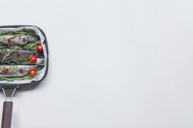 elevated view of fish with rosemary, bay leaves and cherry tomatoes in tray with baking paper on white table clipart