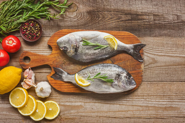 top view of raw fish on wooden board with rosemary and lemon on table