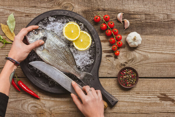 cropped image of tattooed woman cutting raw fish by knife on wooden table