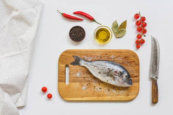 flat lay with uncooked fish on wooden board and various ingredients on white table