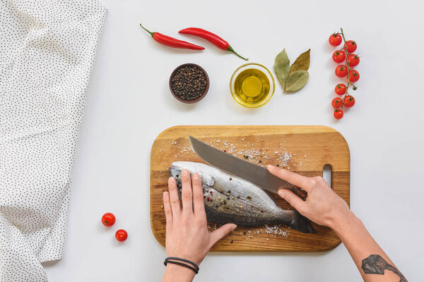cropped image of tattooed woman cutting fish by knife on wooden board near ingredients on table