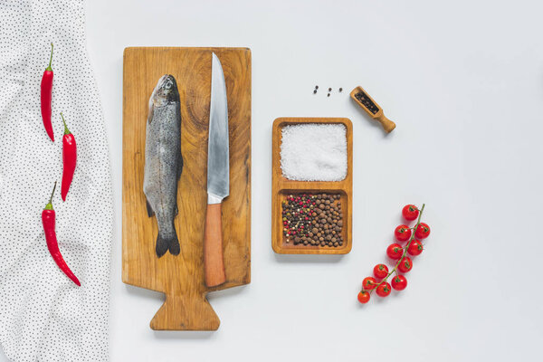 flat lay with knife and uncooked fish on wooden board near ingredients on white table 