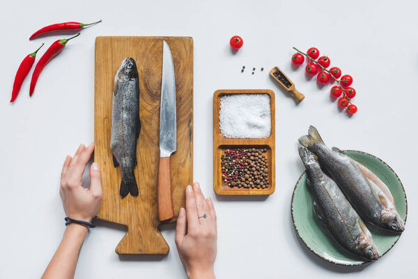 частичный вид женских рук на деревянной доске с ножом и сырой рыбой рядом с ингредиентами на белом столе
