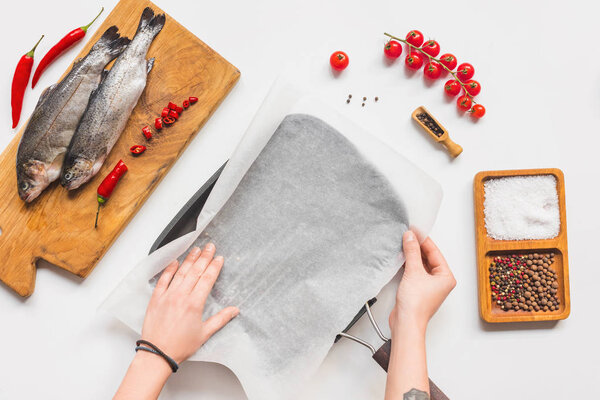 partial view of woman putting baking paper into tray near uncooked fish with ingredients on white table