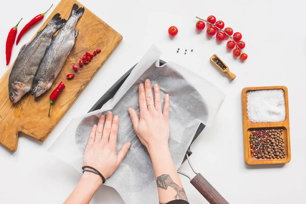 cropped image of woman putting baking paper into tray near uncooked fish with ingredients on white table