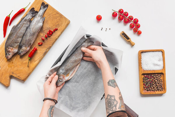 partial view of woman putting uncooked fish in tray with baking paper on white table with ingredients