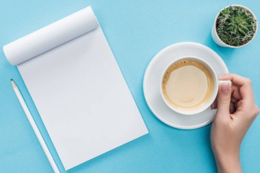 fincan kahve, boş defter ve kalem mavi arka plan üzerinde tutan kişinin kırpılmış görünümü