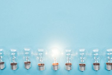 top view of light bulbs in row and one of them glowing on blue background, having new ideas concept clipart