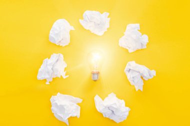glowing light bulb in circle of crumbled paper balls on yellow background, having new ideas concept clipart