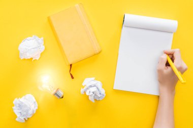 cropped view of woman writing in blank notebook, crumbled paper balls and glowing light bulb on yellow background, having new ideas concept clipart