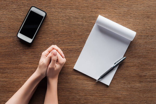 cropped view of person with folded hands, pen, notebook with copy space and smartphone with blank screen on wooden background