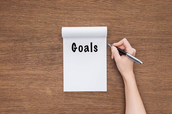 cropped view of woman holding pen and notebook with 'goals' lettering on wooden background 