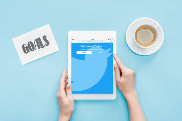 cropped view of female using digital tablet with twitter app on screen, goals lettering on card and coffee on blue background