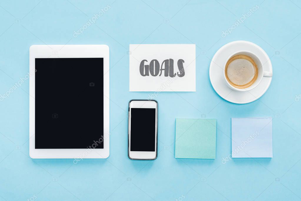 organized flat lay of digital tablet and smartphone with blank screens, sticky notes and 'goals' lettering on card with blue background
