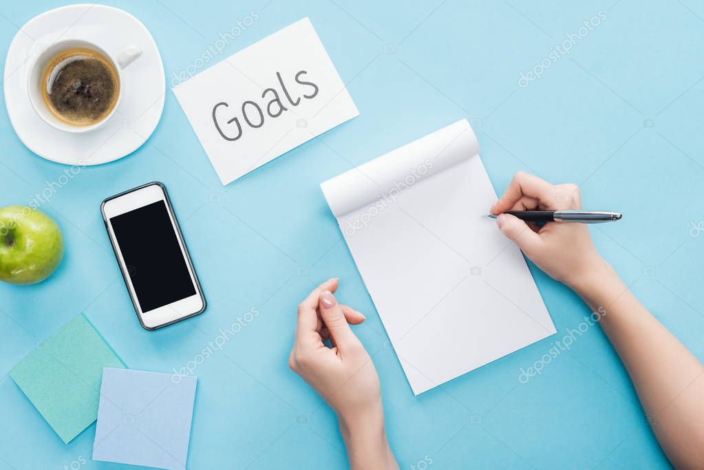 cropped view of woman writing in notebook, lettering 'goals' on card, coffee, sticky notes and smartphone with blank screen on blue background 