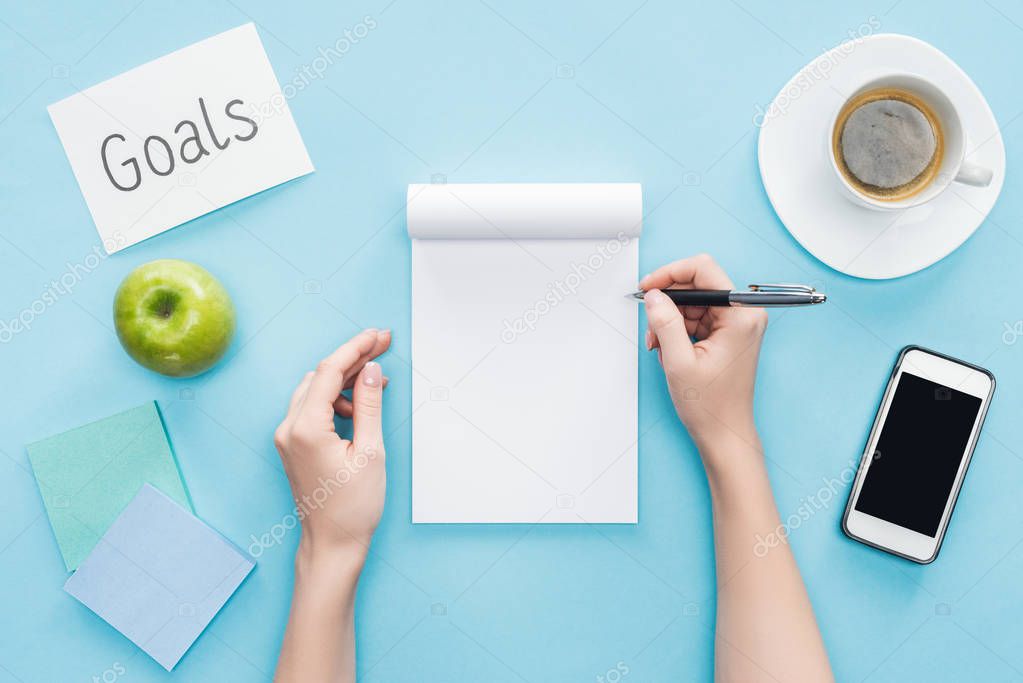 cropped view of woman writing in notebook, lettering 'goals' on card, coffee, sticky notes and smartphone with blank screen on blue background 