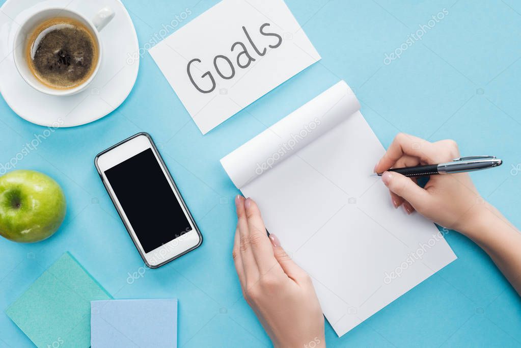 cropped view of woman writing in notebook, lettering 'goals' on card, coffee, sticky notes and smartphone with blank screen on blue background 