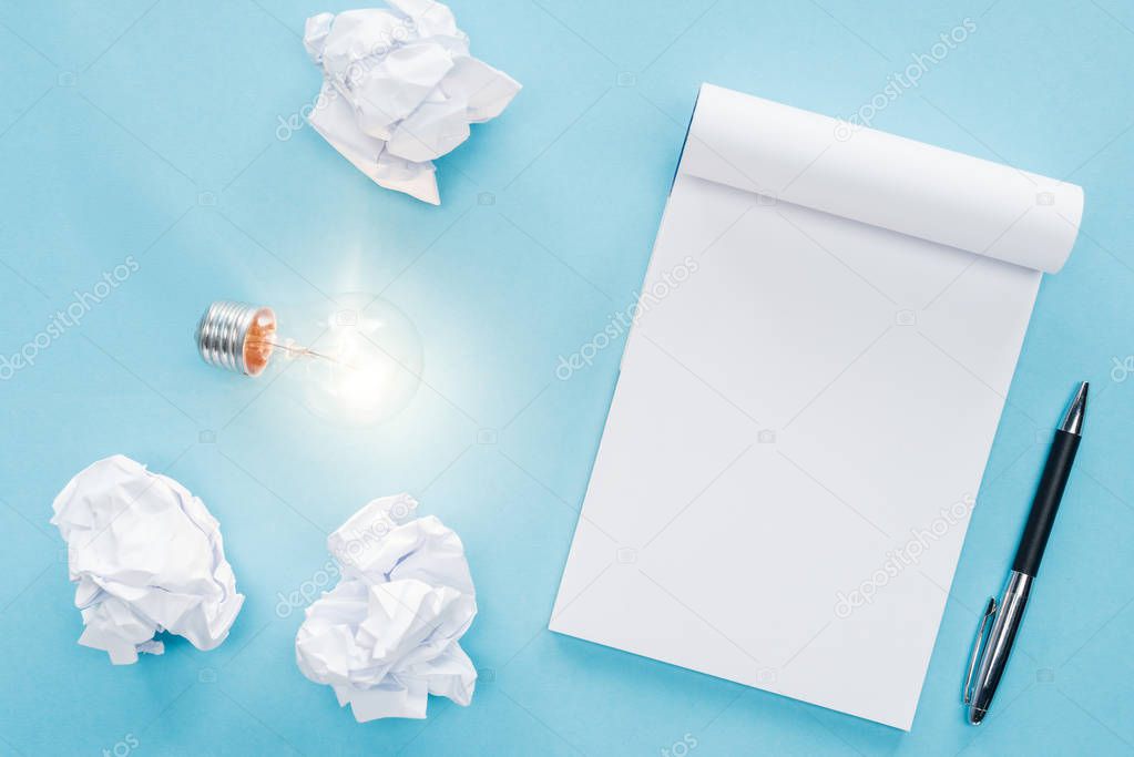 top view of blank notebook with crumbled paper balls and glowing light bulb on blue background, having new ideas concept