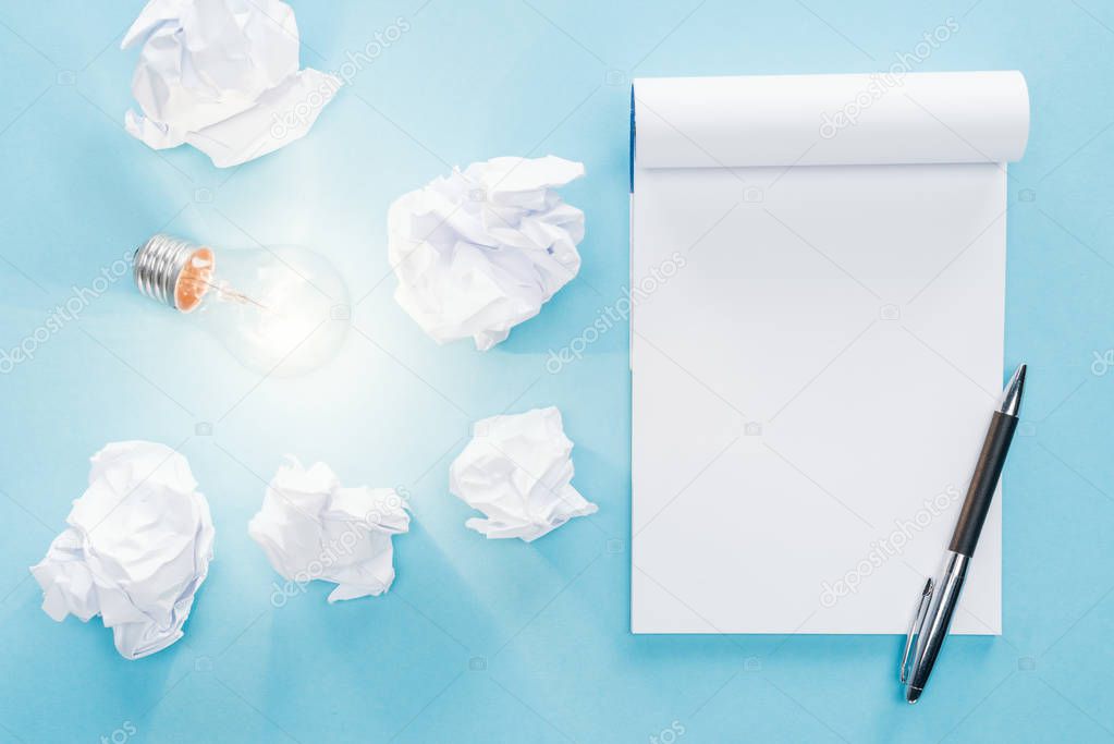 top view of blank notebook with crumbled paper balls and glowing light bulb on blue background, having new ideas concept