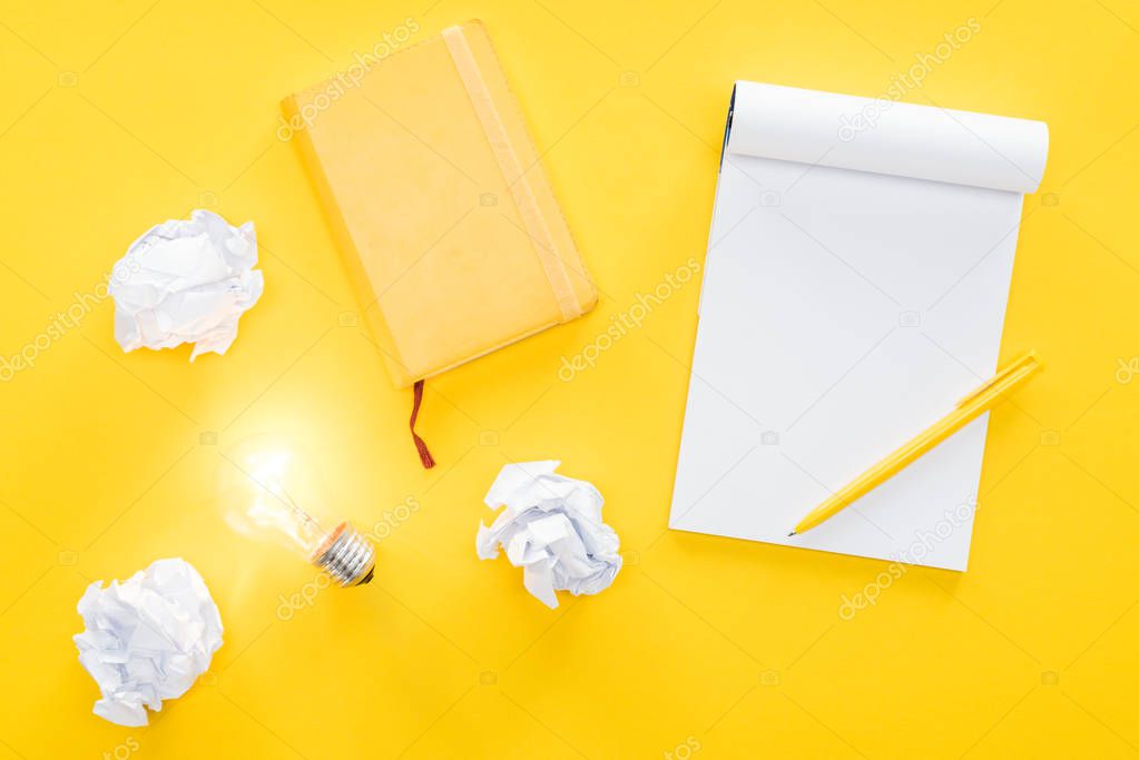 top view of blank notebook, crumbled paper balls and glowing light bulb on yellow background, having new ideas concept