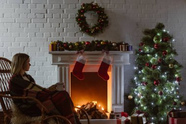  Hasır sallanan sandalyede oturma odasında kahve tutan genç kadın Noel için dekore edilmiştir. 