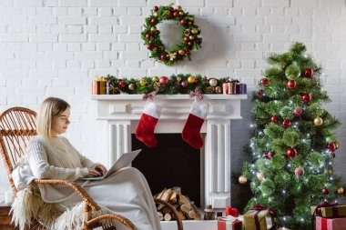  Hasır sallanan sandalyede oturma odasında dizüstü kullanarak genç kadın Noel için dekore edilmiştir. 