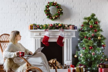 güzel ve genç birbayan hasır sallanan kahve fincanı tutarak ve oturma odasında dizüstü kullanarak sandalye üzerinde Noel için dekore edilmiştir. 