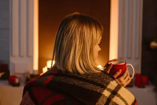 后面的妇女在格子毯子坐在壁炉附近 拿着一杯咖啡在家里 — 免费的图库照片