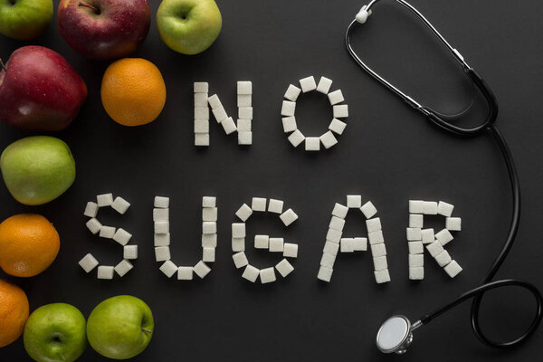 top view of "no sugar" phrase made of sugar cubes with stethoscope and fruits on black background