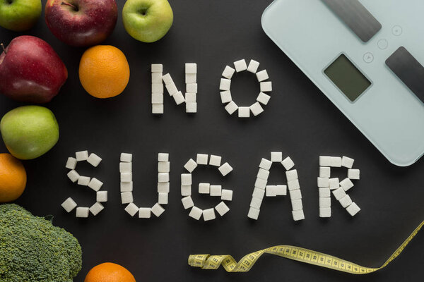 top view of "no sugar" phrase made of cubes among fruits with scales on black 