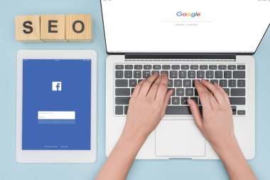 Laptop klavye ile açılan google tarayıcısı dijital tablet yakınındaki açık mavi renkli facebook app ile yazarak kadının üstten görünüm 