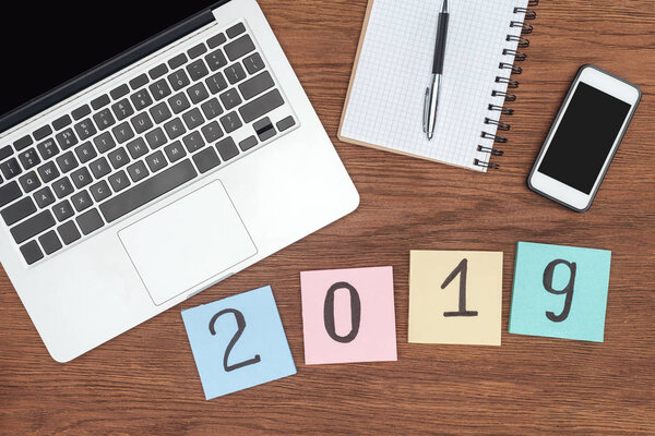 top view of wooden desk with laptop, notebook, smartphone with blank screen and 2019 date made of sticky notes