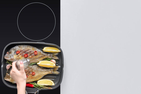 cropped shop of person adding salt while cooking fish with rosemary and lemon on grill 