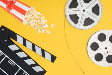 top view of clapperboard, overturned striped bucket with popcorn and film reels with cinema tape isolated on yellow clipart