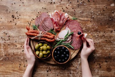 cropped view of  woman taking prosciutto from round cutting board with olives, salami and ham on wooden table with scattered peppercorns clipart