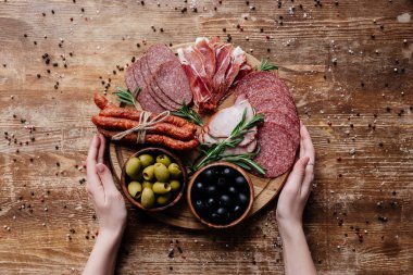 kadın üstten görünüm yuvarlak kesme tahtası zeytinli iki kase içinde tutan eller ve dilimlenmiş salam, jambon ve jambon dağınık peppercorns ahşap tablo
