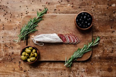 Biberiye, siyah ve yeşil zeytin iki kase ve lezzetli dilimlenmiş salam kesme tahtası üstten görünüm dağınık baharat ahşap tablo