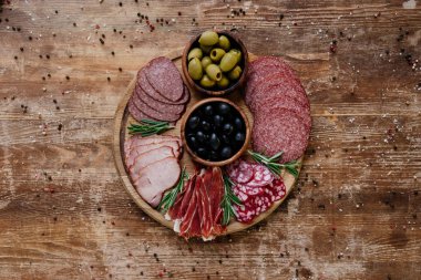 top view of round cutting board with olives and sliced salami, prosciutto and ham on wooden table with scattered spices clipart