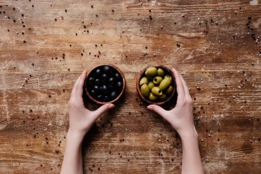 female hands holding two bowls with black and green olives on wooden table clipart