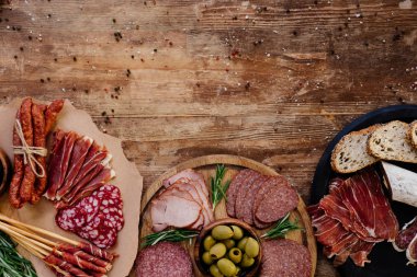 top view of cutting boards with delicious prosciutto, salami, bread, olives, breadsticks, and herbs on wooden table  with scattered spices clipart