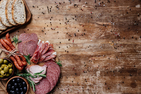 top view of cutting boards with delicious salami, smoked sausages, olives and bread on wooden table with scattered peppercorns