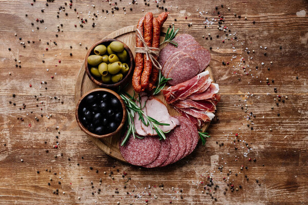 top view of round cutting board with delicious salami, prosciutto and olives in two bowls on wooden table with scattered spices