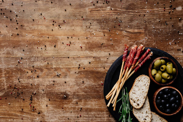 top view of black marble cutting board with olives in bowls, breadsticks, prosciutto, bread and herbs on wooden table with scattered peppercorns