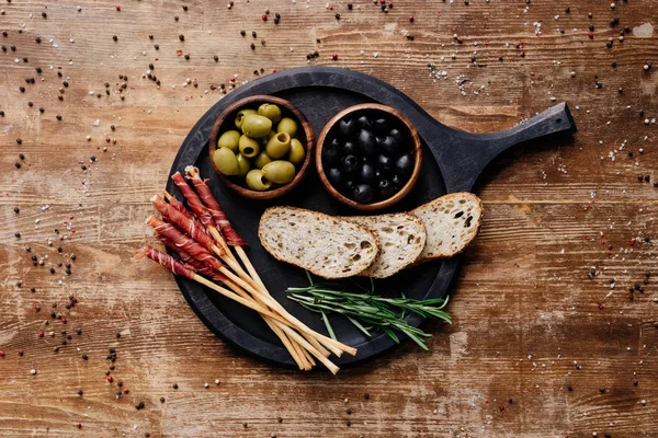 黑色大理石切割板的顶部视图与橄榄 面包棒 意大利饭 面包和迷迭香木桌上的分散的胡椒 — 图库照片