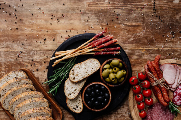 top view of cutting boards with olives, breadsticks, prosciutto, salami, bread, tomatoes and herbs on wooden table