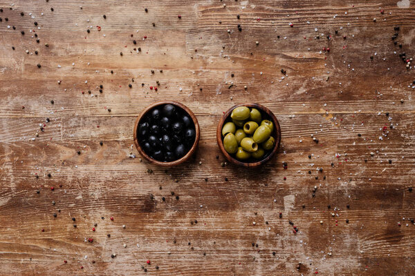 top view of two bowls with black and green olives on wooden table with scattered peppercorns