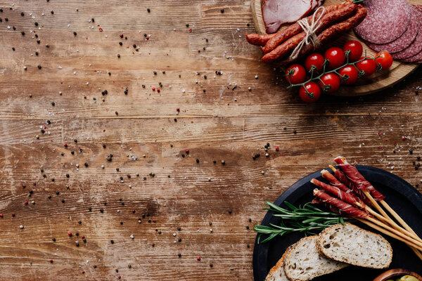 top view of cutting boards with olives in bowls, delicious smoked sausages, salami, bread, tomatoes and herbs on wooden table with scattered spices