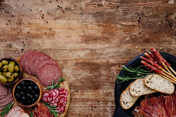 top view of cutting boards with olives, breadsticks, prosciutto, salami, bread and herbs on wooden table